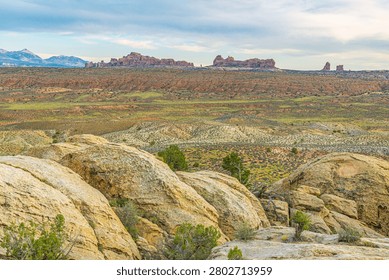 Distant rock formations of the Windows Section rise above a desert valley, viewed past lichen-covered boulders, Arches National Park, Utah.