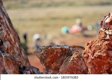 Distant Shot, Blurred Image Of A Family Having Picnic In A Sunny Autumn Day In The Countryside Under Mineral Rich Red Rocky Cliff, Focussed On The Rocks