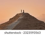Distant side view of a couple of people standing atop a rocky hill with reddish orange skies in the background.