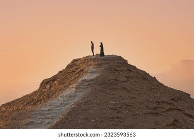 Distant side view of a couple of people standing atop a rocky hill with reddish orange skies in the background. - Powered by Shutterstock