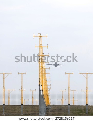 A distant view of an airplane taking off, flying over a series of yellow runway lights on a cloudy, overcast day. The image highlights the infrastructure of airport operations and the dynamics of flight departure.