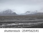 Distant view of a glacier nestled between two snow-dusted mountains in Iceland. The foreground showcases a vast expanse of volcanic terrain, emphasizing the rugged beauty of the landscape