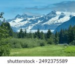 Distant view of Mendenhall Glacier (middle ground) and mountains across green meadow with a creek (bottom right) and conifers along Mendenhall Valley on the outskirts of Juneau, Alaska, in mid June