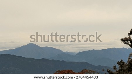 Distant view of Mount Fuji summit covered with snow appearing through clouds behind mountain ranges.