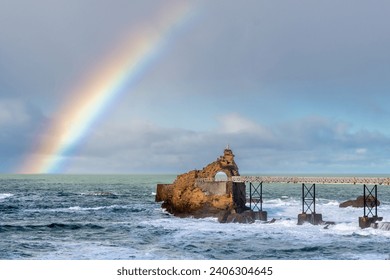 Distant view of the Rocher de la Vierge (meaning 'Rock of the Virgin') in Biarritz, France, after the rain, with a beautiful rainbow over the Atlantic Ocean