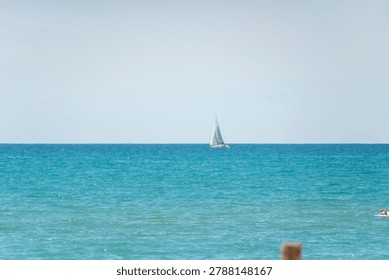 Distant view of a white sailboat with sails up navigating the blue Mediterranean Sea on a bright sunny day under a clear sky.