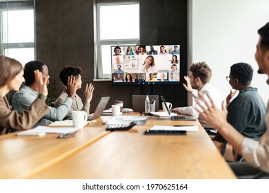 Distant Virtual Meeting Concept. Diverse Business People Making Online Video Call With Colleagues, Sitting At Desk In Boardroom, Waving Hands At Tv Screen At Office, Talking On Web With Remote Workers