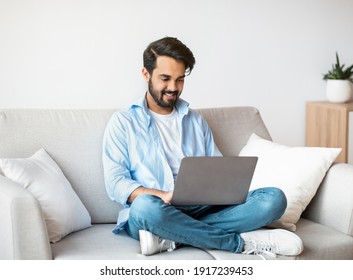 Distant Work. Happy Arab Freelancer Man Using Laptop Computer At Home, Typing On Keyboard, Young Eastern Guy Sitting On Sofa In Living Room, Working Remotely On Online Project, Copy Space