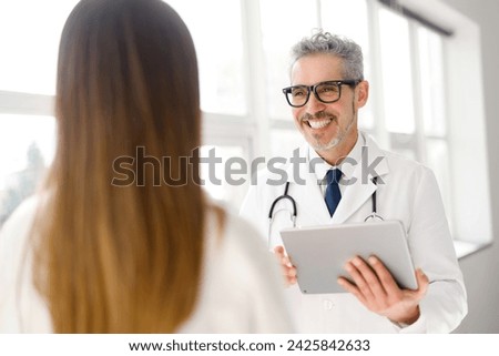 A distinguished doctor with grey hair is shown holding a tablet and engaging with a young female patient in a bright, modern clinic