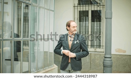 Distinguished man with beard fastening suit jacket by a reflective glass building in an urban setting.