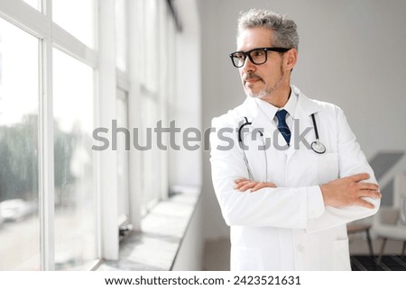 A distinguished mature doctor with grey hair looks thoughtfully out of the window in his clinic, suggesting a moment of reflection amid a busy healthcare practice
