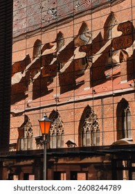 Distorted reflection of old red brick church with arched windows and tiled roof on modern glass building facade with orange street lamp in foreground. Warped mirror, red cathedral, orange streetlight