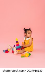 Distracted Asian Toddler Girl In Yellow Dress Playing Building Blocks On Pink