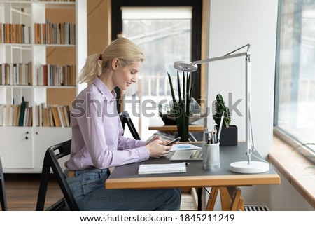 Distracted from computer work young addicted to technology beautiful woman using mobile phone applications, chatting in social networks, playing game or web surfing, sitting alone at workplace.