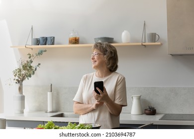 Distracted from cooking smiling dreamy older mature retired woman in eyeglasses holding cellphone in hands, looking in distance, having fun enjoying domestic hobby activity alone in modern kitchen.