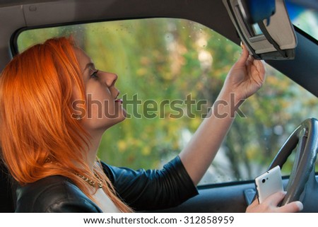 Distracted driver. Busy woman looking at mirror, using phone while driving her car