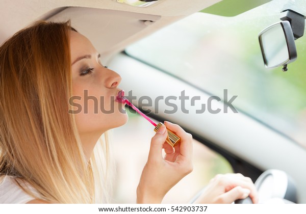 Distracted driver. Young attractive woman looking in\
rear view mirror painting her lips doing applying make up while\
driving the car.