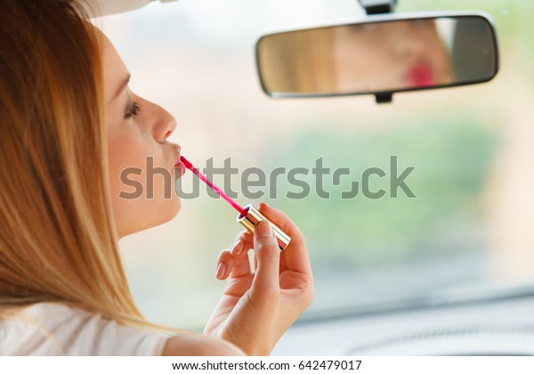 Distracted driver. Young attractive woman looking in\
rear view mirror painting her lips doing applying make up while\
driving the car.