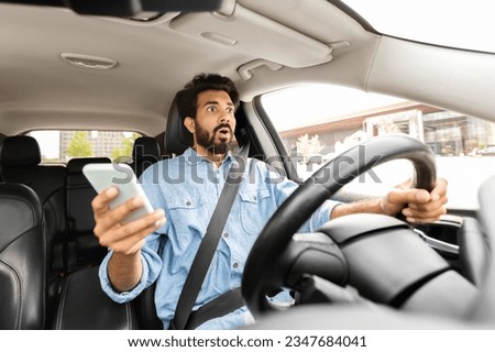 Distracted driving concept. Shocked scared handsome young indian man in casual outfit driver using smartphone while driving auto, got car accident. Guy holding phone in his hand, crushed automobile