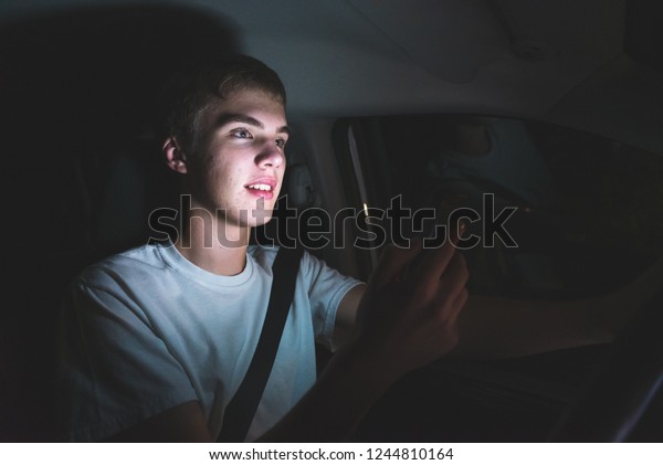 Distracted teenager driving a car with his cell\
phone in his hand. The light from the screen of the phone is\
illuminating his\
face.