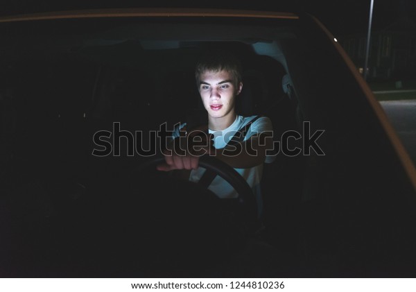 Distracted teenager driving a car with his cell\
phone in his hand. The light from the screen of the phone is\
illuminating his\
face.