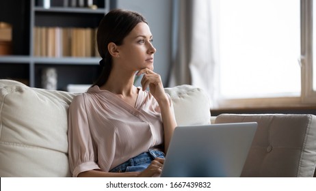 Отвлеченная от работы взволнованная молодая женщина сидит на диване с ноутбуком, думая о проблемах. Задумчивая немотивированная дама смотрит в окно, чувствует нехватку энергии, выполняет удаленные внештатные задания дома.