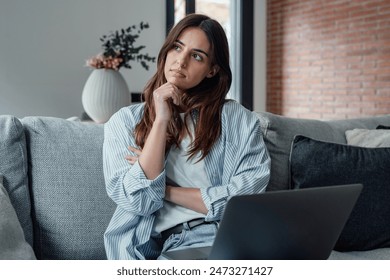 Distracted from work worried young woman sitting on couch with laptop, thinking of problems. Pensive unmotivated lady looking at window, feeling lack of energy, doing remote freelance tasks at home.