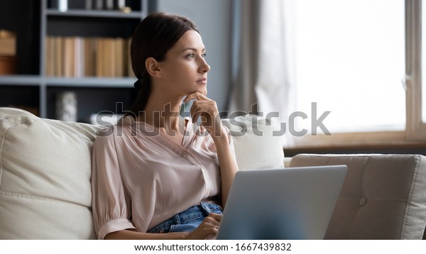 Distracted from work worried young woman sitting on\
couch with laptop, thinking of problems. Pensive unmotivated lady\
looking at window, feeling lack of energy, doing remote freelance\
tasks at home.