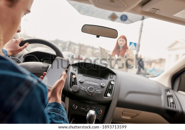 Distracted young male driver looking at the screen\
of his mobile phone while running over a pedestrian. Technology and\
transportation concept. Selective focus on hand with smartphone.\
Horizontal shot