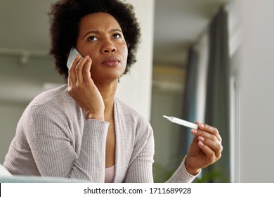 Distraught African American Woman Communicating Over Mobile Phone While Measuring Her Temperature At Home. 