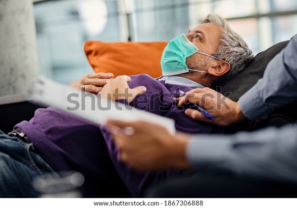 Distraught man lying down on\
psychiatrist\'s couch and wearing a face mask due to coronavirus\
pandemic.