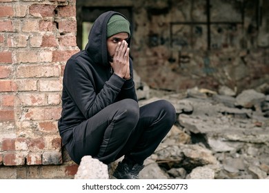 Distraught Young Black Refugee Man In Black Outfit Crouching At Ruined Building And Covering Face With Hands