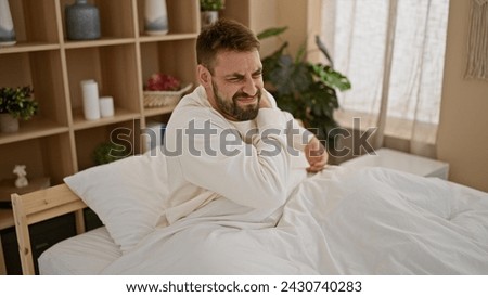 Distraught young hispanic man suffering from severe back injury, touching spinal column in agony while awake on his bedroom bed, morning light casting a shadow on his worried expression.