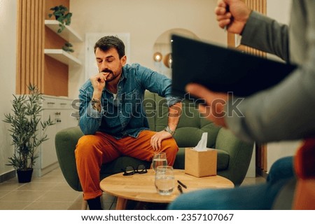 A distressed client is sitting on the sofa and contemplating during his session with a shrink in the office. A client is thinking while a therapist is asking questions and writing notes in a textbook.