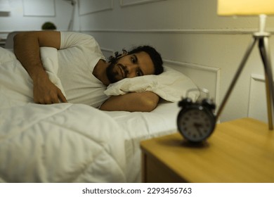 Distressed hispanic man lying awake late at night and watching the alarm clock while suffering from insomnia