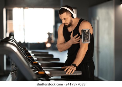 Distressed man experiencing sudden heartache while working out on treadmill, young male athlete showing signs of discomfort and pain during training in gym, touching chest area, copy space - Powered by Shutterstock