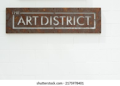 A Distressed Metal Sign With The Text Art District Cutout. The Sign Is Hanging On A White Brick Wall. The Signage Is For A Public Space For Galleries, Theatres And Modern Performances.  