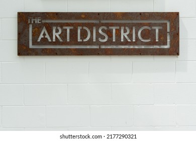 A Distressed Metal Sign With The Text Art District Cutout. The Sign Is Hanging On A White Brick Wall. The Signage Is For A Public Space For Galleries, Theatres And Modern Performances.  