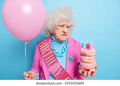 Distressed Unhappy Elderly Woman Being Upset About Getting Older Holds Pile Of Glazed Doughnuts Celebrates Birthday Alone Feels Lonely Dressed In Fashionable Clothes Poses With Balloon Indoor