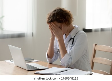 Distressed woman doctor in white medical uniform sit at desk in hospital feel stressed upset with bad news on laptop, unhappy tired female nurse or GP suffer from overwork or migraine at workplace