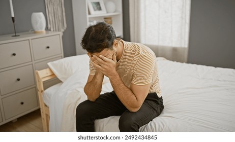 A distressed young hispanic man sits on a bed in a modern bedroom, covering his face with his hands.