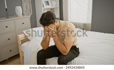 A distressed young hispanic man sits on a bed in a modern bedroom, covering his face with his hands.