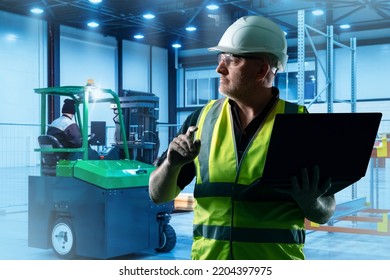 Distribution Center Manager. Man In Yellow Vest Looks Away. Manager With Laptop Inside Distribution Warehouse. Forklift Behind Man. Experienced Guy Manages Work Of Distribution Center