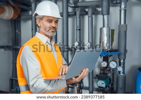 District heating plant interior and experienced technician standing by pipeline energy source using laptop
