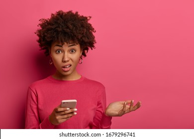 Distrubed dark skinned woman has problematic concerned face expression, holds smartphone, raises palm, cannot understand whats wrong, strange meaning of message, shrugs shoulders, stands indoor - Shutterstock ID 1679296741