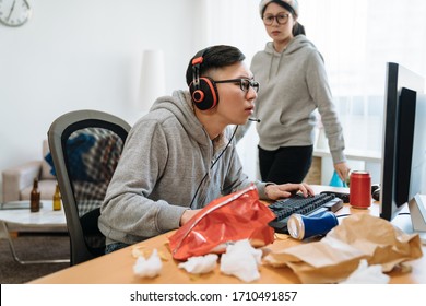 Distrustful Girlfriend Annoyed With Boyfriend Technology Addiction. Bokeh Angry Young Asian Woman Staring At Guy And Walking Clean Up While Man Playing On Computer Online Game In Messy Room At Home