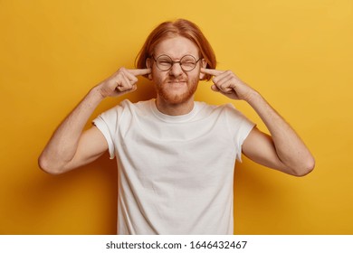 Disturbed ginger man plugs ears, ignores loud music, smirks face, hears unpleasant annoying sound, dissatisfied by noise, wears casual clothing, isolated on yellow wall. Noisy neighbours is problem