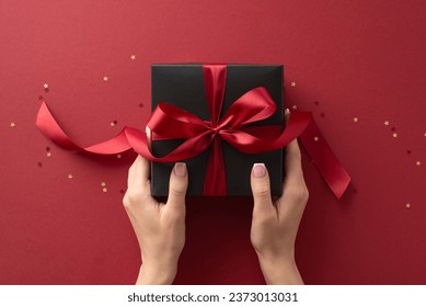 Dive into holiday magic! First person top view of hands reach for lavish black giftbox featuring ribbon bow, nestled among shimmering confetti on marsala backdrop. Perfect for Black Friday marketing