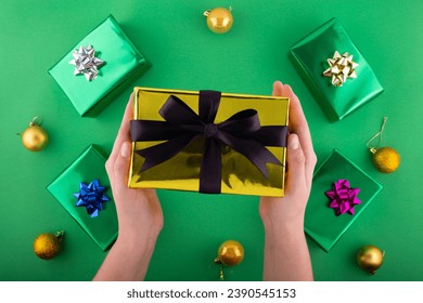 Dive into holiday magic! First person top view of hands reach for lavish golden giftbox featuring ribbon bow near green gift boxes surprise on green backdrop. Perfect for Black Friday marketing
