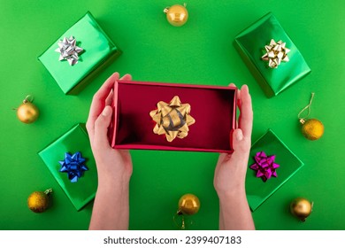 Dive into holiday magic! First person top view of hands reach for lavish red giftbox featuring ribbon bow near green gift boxes surprise on green backdrop. Perfect for Black Friday marketing